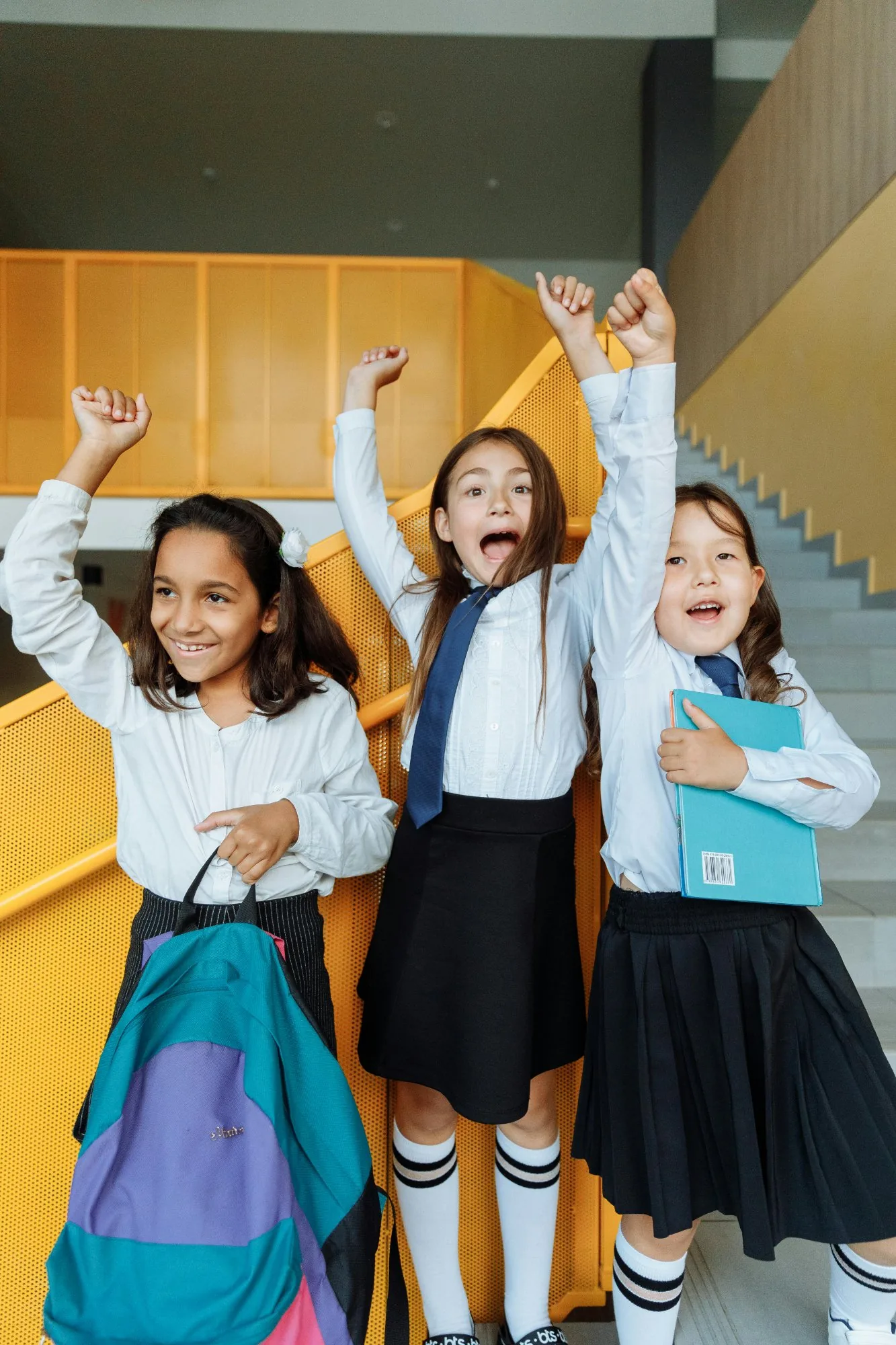 Three excited primary school girls celebrating with arms raised in school corridor